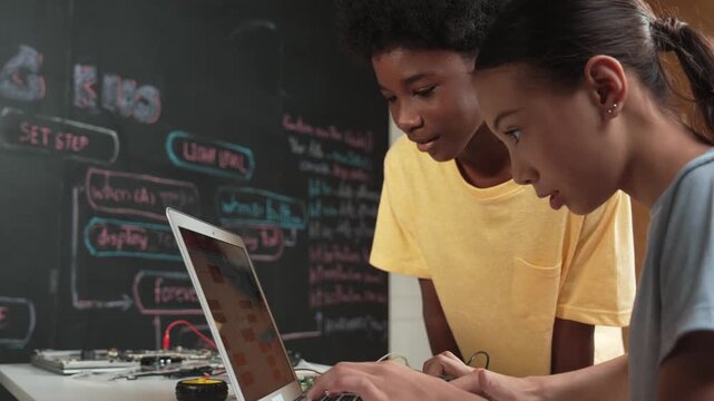 Happy african boy pointing at laptop screen while cute girl typing laptop coding or programming engineering system. Teacher teaching program while holding laptop at STEM technology class. Edification