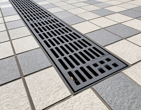 Black metal drainage grate installed in a pedestrian walkway with a checkered pattern of light grey and dark grey stone tiles in an outdoor urban public environment setting