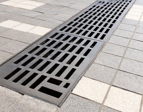 Black metal drainage grate embedded in a pedestrian walkway paved with alternating light and dark grey stone tiles captured in a high angle perspective during the daytime