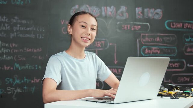 Smart girl programming system by using laptop at blackboard. high school student writing engineering code or prompt while looking at camera at STEM technology class. Creative education. Edification