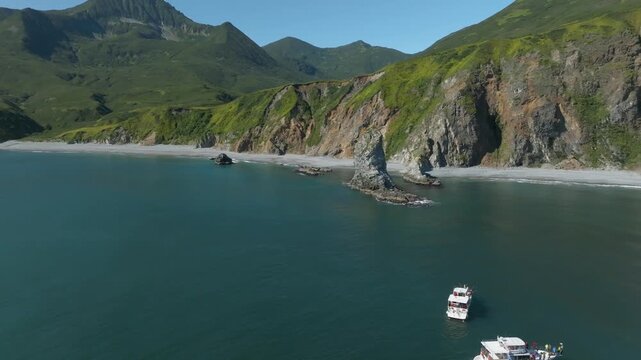 Drone footage of rugged Avacha Bay shoreline with steep cliffs, green volcanic hills, quiet sea and small tour boats near the coast, Kamchatka Krai, Russia