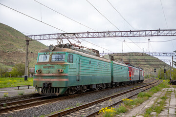 Fototapeta premium Electric freight locomotive, a vl10 model, waiting on tracks at a provincial railway station in armenia, with an overhead line system and hills in the background on an overcast day