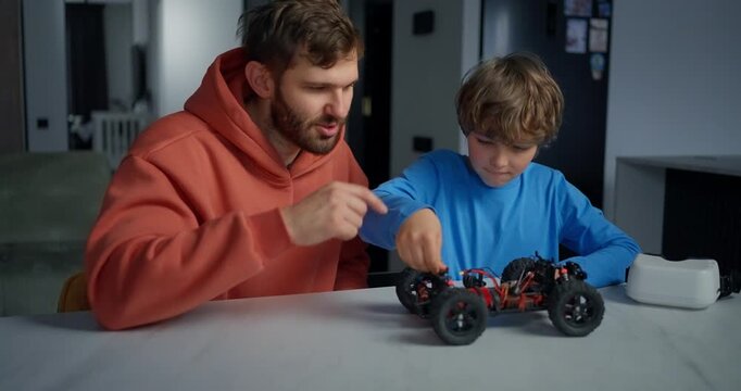 Father and son work together on a small remote control car at a kitchen table, sharing a focused hands-on lesson in mechanics and play
