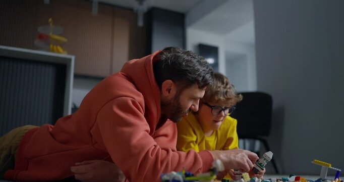 Father and young son lying on floor building colorful toys together, sharing focused creative play and bonding in a cozy modern home