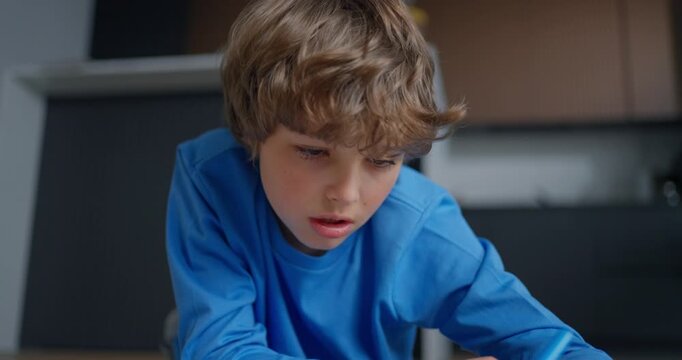 Focused young boy in a blue shirt writing with a pen on paper at a home table, concentrating on homework in a softly blurred indoor setting
