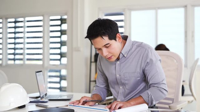 Young asian civil engineer studying a blueprint floor plan and  working with documents, drawings and building models.