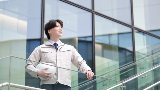 ビルの前でヘルメットを持ちながら未来を見つめるハンサムな若手作業員のポートレート　施工管理、ゼネコンの都市開発、メンテナンスなどのイメージの動画