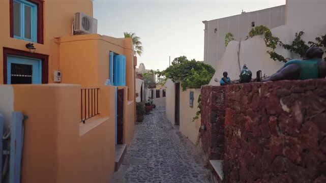 Picturesque cobblestone alley meanders through charming Santorini, Greece island village featuring warm orange stucco buildings with vibrant turquoise shutters, red stone walls, lush green vegetation.
