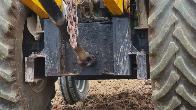 Close view of tractor PTO shaft rotating to power auger drill during ground drilling, mechanical linkage and wheels visible in a rural agricultural setting