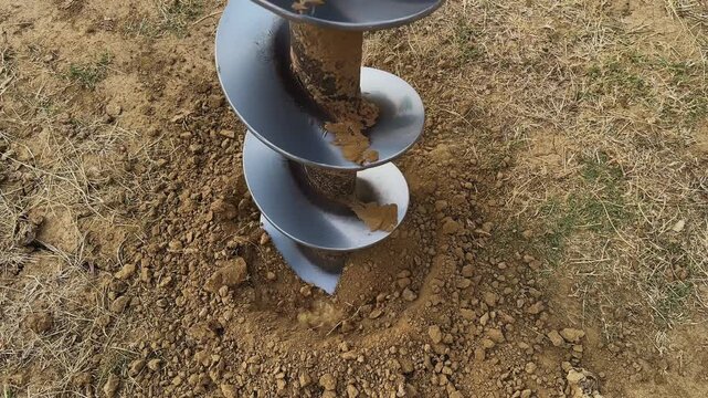 Close view of auger drill bit rotating into soil creating hole for fencing pole, loose earth and gravel surrounding in a rural outdoor construction setting