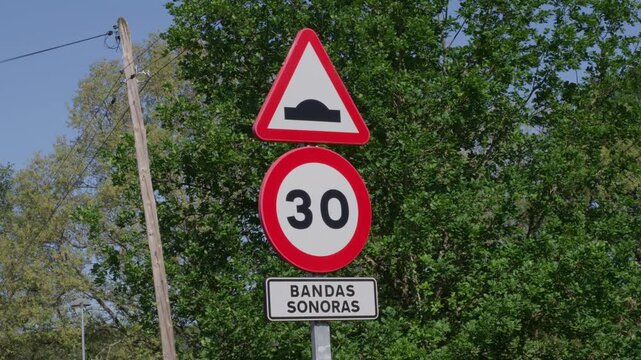 Clear view of speed bump and 30 kmh limit road sign labeled bandas sonoras by green trees, sunlight, and blue sky. Nobody, static shot.
