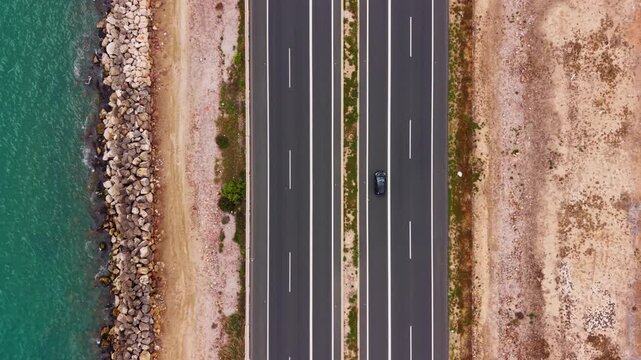 Top-down view of a multi-lane coastal highway with cars driving alongside turquoise ocean waters, rocky shoreline, and arid sandy terrain on the opposite side