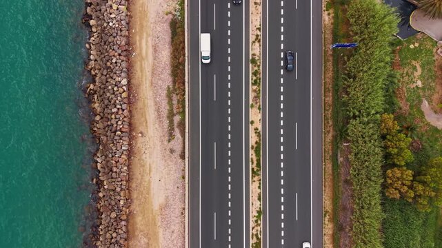 Top-down view of a multi-lane coastal road with cars driving alongside turquoise water, rocky shoreline, sandy strip, and lush green trees on the opposite side