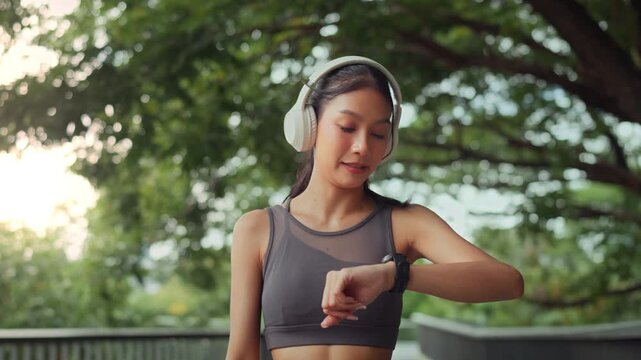 Active young Asian woman wearing headphones and checking her smartwatch during an outdoor workout in a lush green park. Focusing on fitness tracking and healthy lifestyle goals.