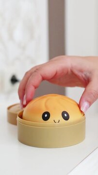 hand lifting yellow kawaii squishy dumpling toy with smiley face out of open plastic steamer box on white table surface close-up