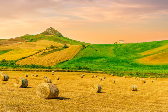 scenic agricultural landscape of summer season golden field with hay stacks during sunset or sunrise with green rural hills and beautiful mountains and clouds in evening sky on background