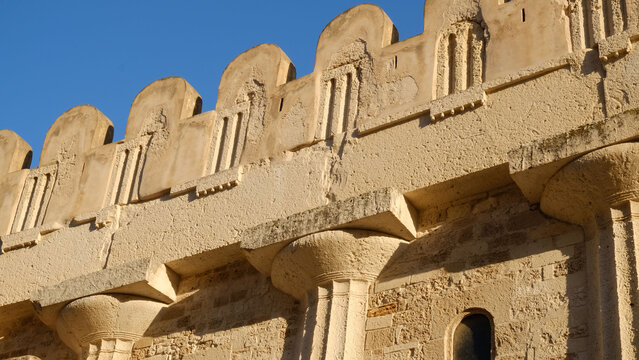 Historic architectural facade with crenellated parapet, carved arches, and sunlit stone texture against a blue sky. For heritage and travel concepts.