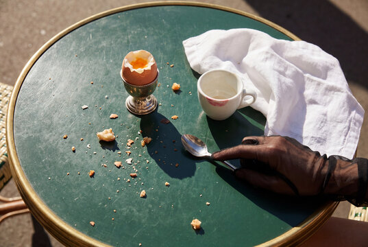 A gloved hand rests on a green bistro table next to a half-eaten soft-boiled egg, an empty cup with a lipstick mark, a silver spoon, a napkin, and bread crumbs.