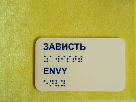 Yellow plastic accessibility plate displays word envy in english and russian cyrillic with raised tactile braille dots for visually impaired people on vibrant textured background