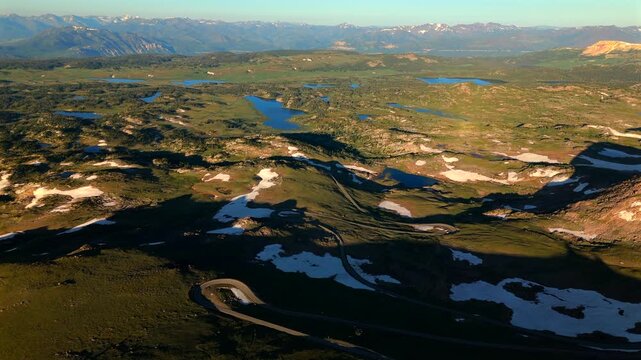 Cinematic Aerial of Beartooth Highway in Montana winds through stunning alpine terrain with scattered lakes and snow patches. Golden hour light illuminates green valleys and distant mountain peaks