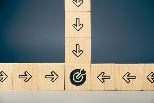 Wooden blocks with arrows moving from multiple directions toward a central target symbol, representing diverse strategies, teamwork, alignment, and different paths leading to the same business goal
