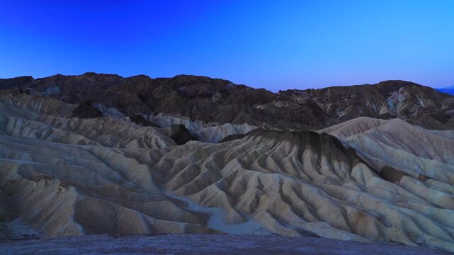 Dramatic twilight view of eroded badlands at Zabriskie Point in Death Valley National Park. Golden sedimentary formations create intricate patterns beneath deep blue sky during dusk.