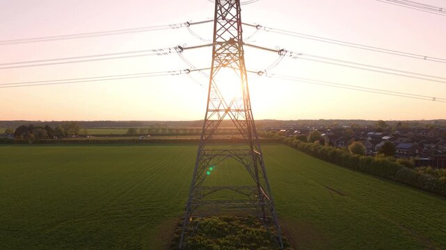 Aerial view of a high voltage electricity pylon standing in a green field at sunset with residential houses in the background in Armthorpe, Doncaster, England, United Kingdom.