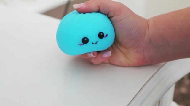 hand tightly gripping and squeezing soft blue kawaii antistress squishy dumpling toy with cute winking smiley face on white table