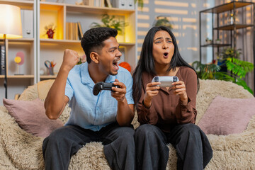 Indian couple man and woman sitting on home sofa play video games with wireless gamepads and smile. Young family man and woman sitting on home sofa enjoy fun gaming and home entertainment with smiles. © Andrii Iemelianenko