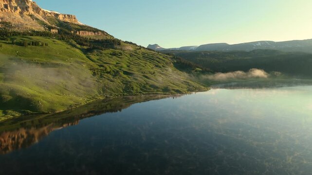 Aerial footage captures a serene mountain lake along Beartooth Highway in Montana during early morning. Mist rises from calm waters reflecting green hillsides and snow-capped peaks.