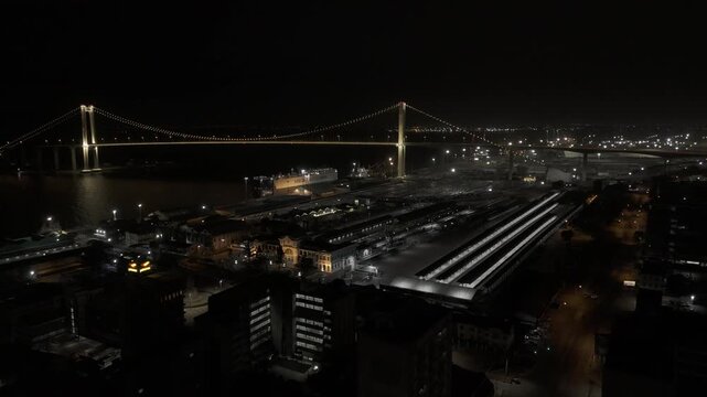 Drone flies over train station toward large suspension bridge lit up at night over the Mbuluzi River in Maputo, Mozambique