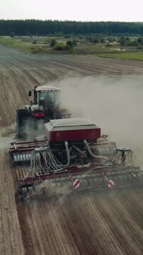 Vertical drone shot of a heavy tractor pulling a modern sowing machine through a dry field. Clouds of dust rising as the agricultural machinery performs spring or autumn planting on a large farm