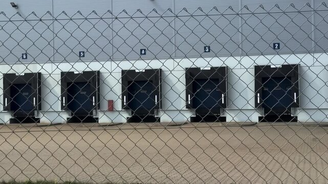 Row of empty loading bays at a large industrial distribution center or warehouse facility viewed through a chain link fence, representing global logistics, supply chain, and freight transportation