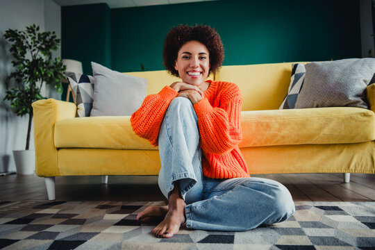 Smiling woman in orange sweater sitting relaxed on the floor in a bright living room with yellow sofa and cozy modern decor