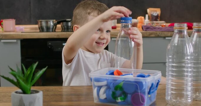Happy boy places blue cap on clear bottle above box filled with sorted lids. Child turns small closure by hand completing bottle handling task during recycling game