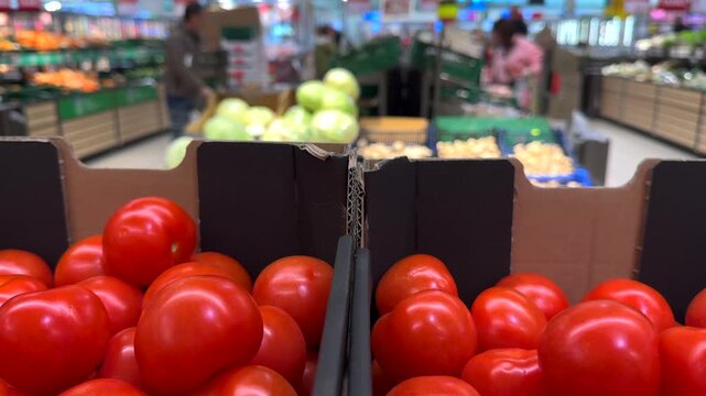 Close up view of fresh red tomatoes in cardboard boxes at a grocery store, with blurred customers and produce aisles in the background representing the daily shopping experience