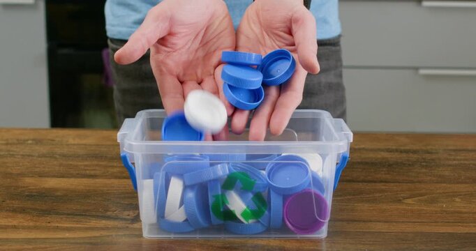 Adult man holds blue plastic caps above clear recycling container on wooden table. Person gathers sorted lids lowering batch into box for household recycling task