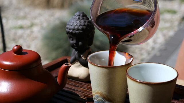 Traditional chinese gongfu tea ceremony in a peaceful garden setting, with black puerh tea pouring from a glass pitcher into ceramic cups next to a clay teapot and a buddha statue