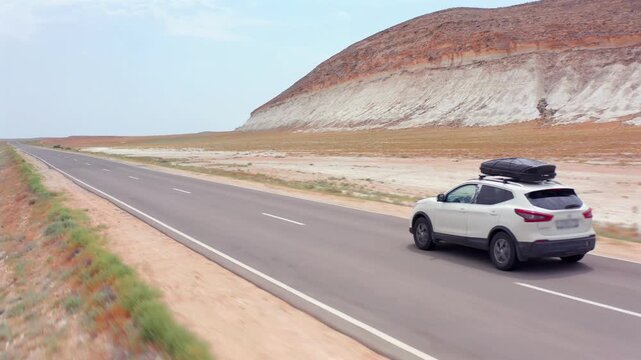 aerial camera tracking car driving sideways along asphalt road, white limestone cliff in Mangystau Kazakhstan, lateral motion across desert terrain with layered hill background showing travel movement