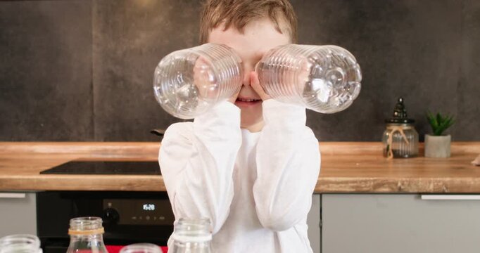 Smiling boy lifts pair of empty bottles looking through transparent plastic like toy binoculars. Child holds clear containers near face playing during recycling break