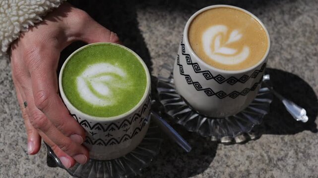 Female hands grabbing a cup of hot matcha green tea latte with beautiful foam art, leaving a cappuccino with a rosetta design on the stone table of an outdoor cafe on a sunny day