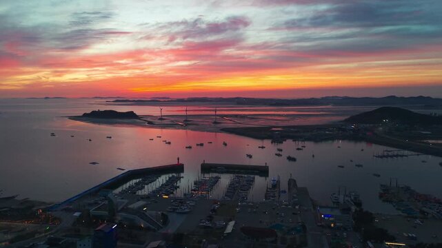 Aerial descending forward drone shot over silhouetted fishing boats toward Nue Island wind turbines at vivid orange post-sunset with Jeongok Marina and calm Korean tidal waters below
