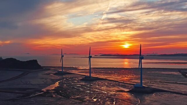 Stationary Three wind turbines on Nue Island tidal sea road as vivid orange sunset blazes over wet mudflats in South Korea - Drone parallax orbits