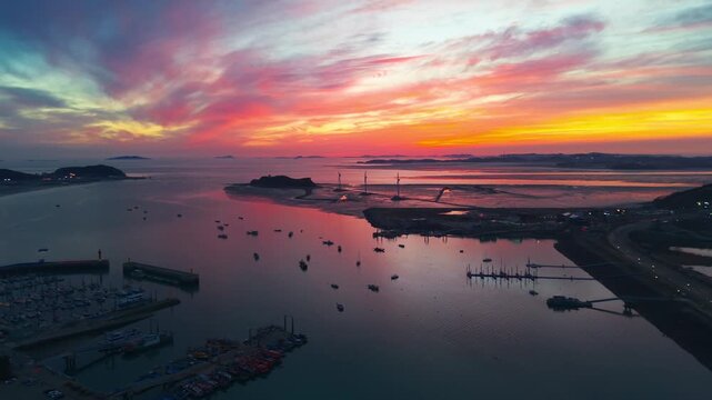 Aerial parallax of Jeongok Marina with silhouetted Nue Island wind turbines and fishing boats reflected in vivid orange and gold sunset sky over calm Korean tidal waters