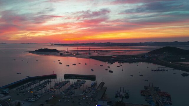 Silhouetted Jeongok Marina, Nue Island wind turbines and Tando Sea Road at soft orange post-sunset afterglow with calm Korean tidal waters reflecting pastel sky - Aerial parallax drone shot