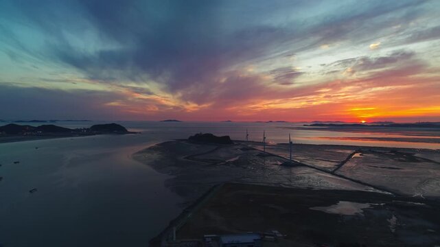 High drone aerial reveals sweeping panorama of Nue Island sea road, coastal village and mudflats under dramatic vivid sunset sky in South Korea