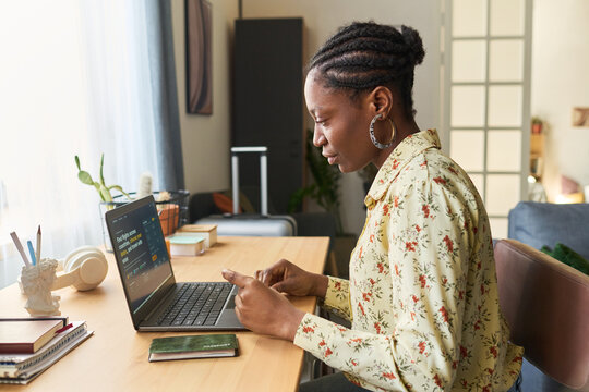 Young African American woman checking smartphone beside laptop and passport, preparing for trip and managing travel booking. Useful for tourism marketing, vacation planning, remote booking