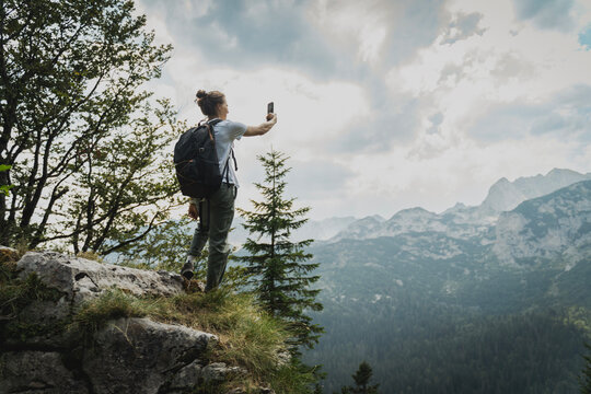 Young woman standing on a mountain peak with a scenic view and holding a mobile phone to take a selfie or photo during a summer hike.