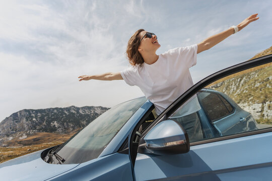 Young happy woman leaning out of a car window with her arms outstretched against the blue sky and mountain landscape, enjoying freedom and summer road trip.