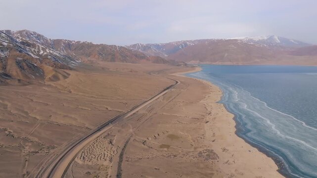 Drone footage of the Orto-Tokoy Reservoir with a winding shoreline road, frozen water edge and barren steppe landscape, framed by snowcapped mountains, Kyrgyzstan